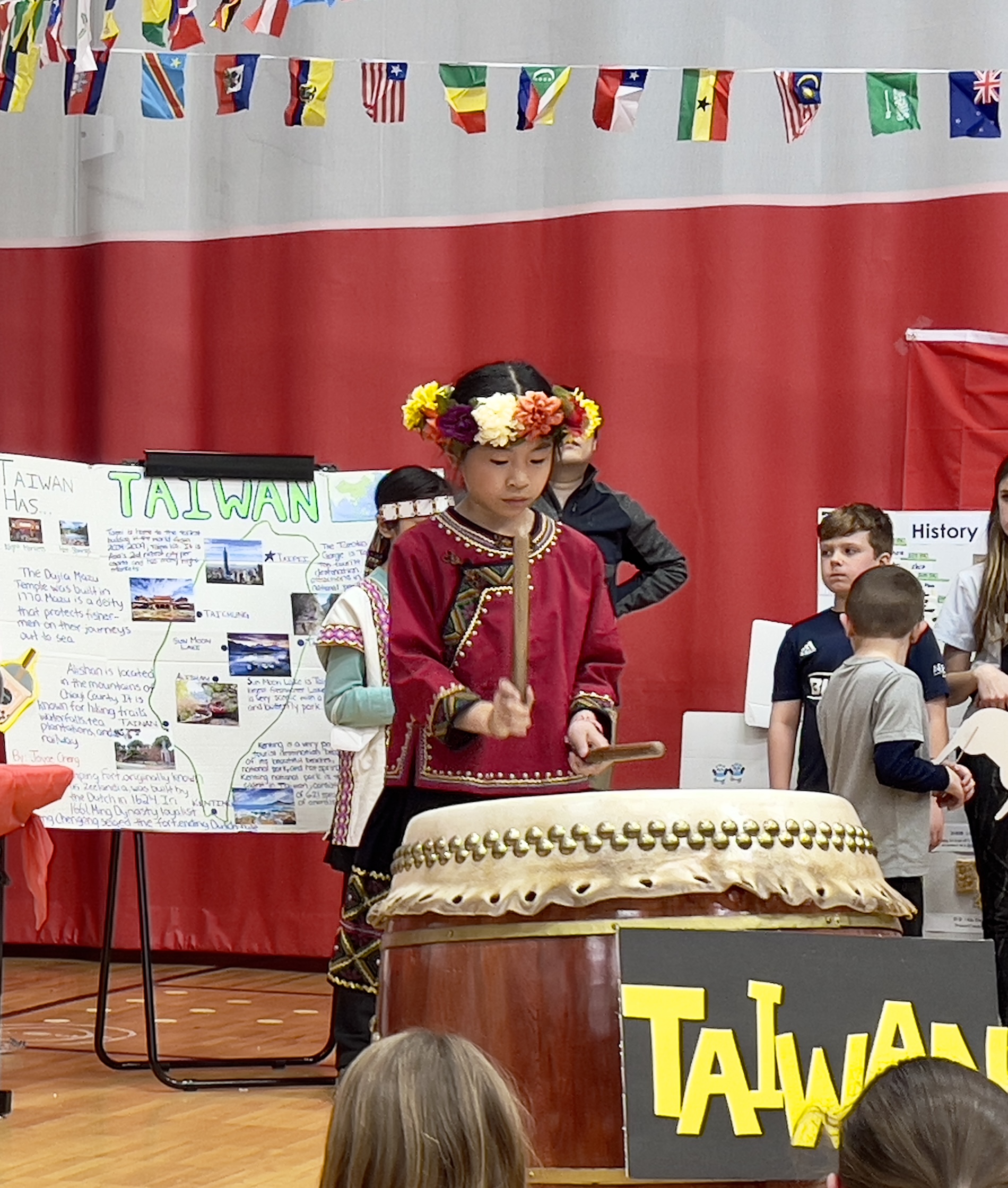 A student wearing traditional clothes from their culture and flowers in their hair plays a large drum.