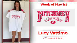 Student stands in front of a while wall holding a white t-shirt with red text that reads: Dutchmen of the Week