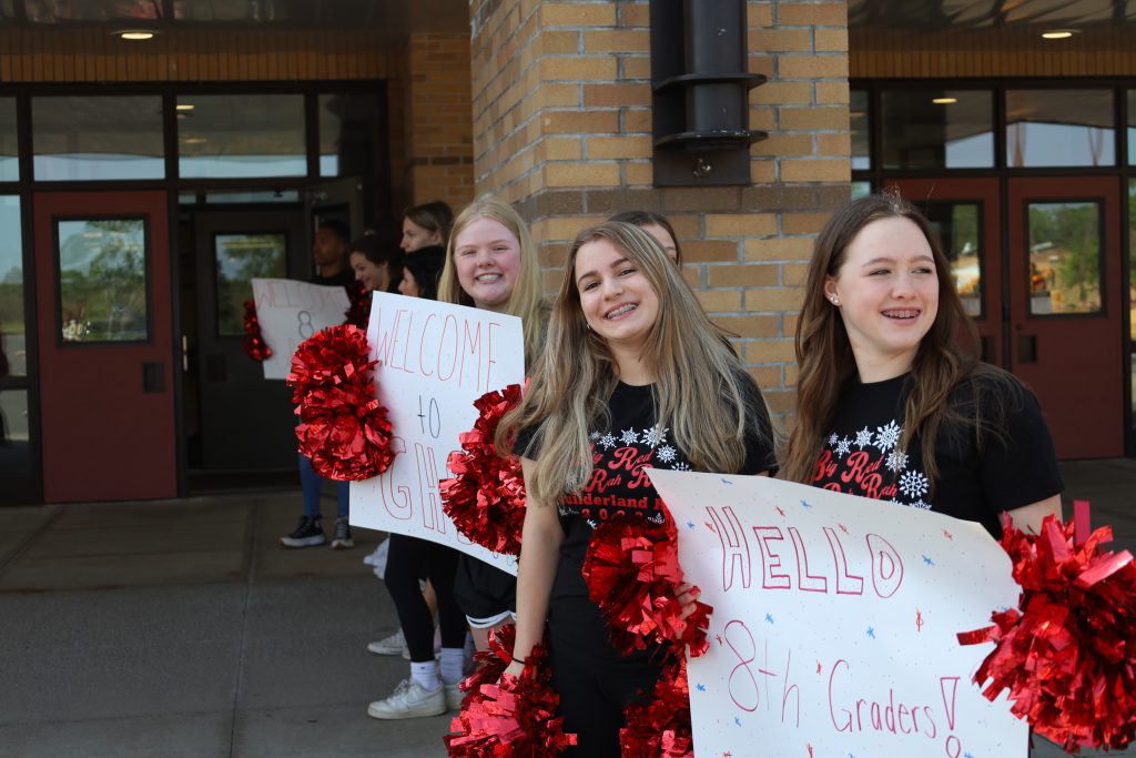 Students are outside, in front of GHS. They are standing in a line, holding signs that read: hello, 8th graders!. They are holding red pom poms.