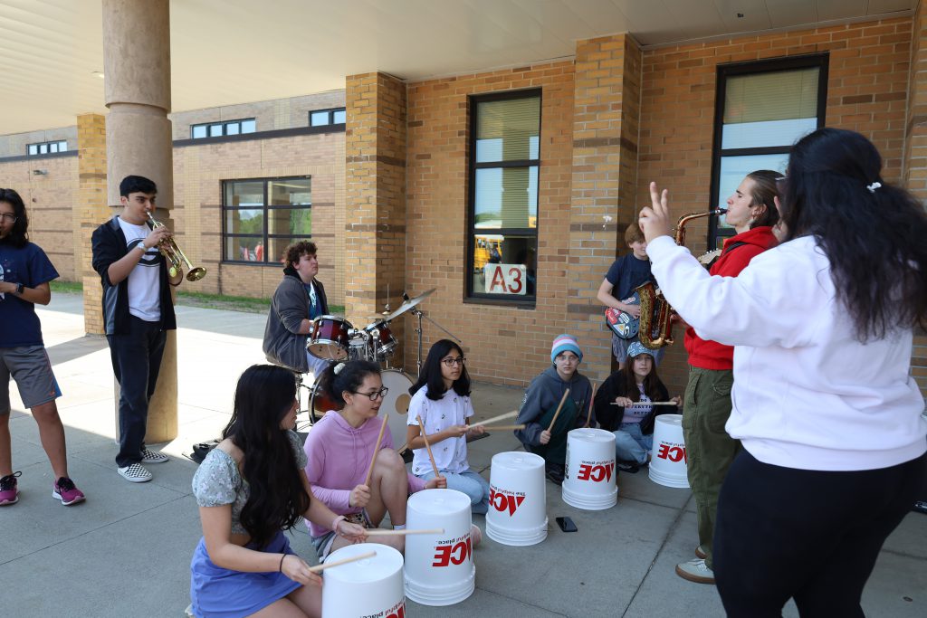 Students are outside, in the front of GHS. Some students are playing musical instruments and others are using plastic buckets for drums.