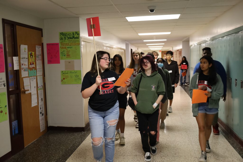 Students walk down the high school hallway. One is leading the others, holding a sign and reading from a paper.