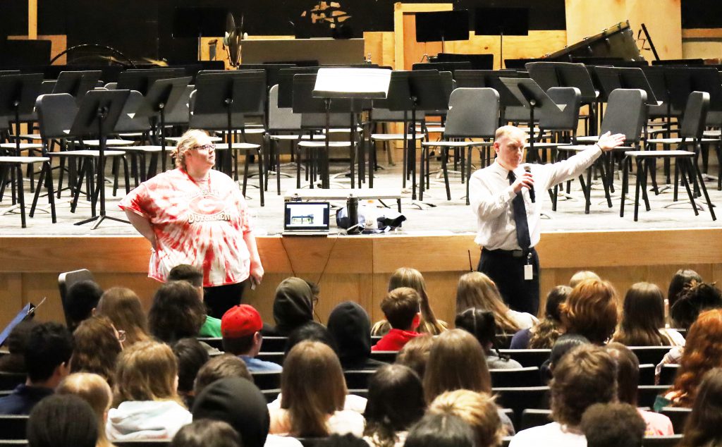 A man stands in front of students seated in an auditorium. He is speaking into a microphone while gesturing.