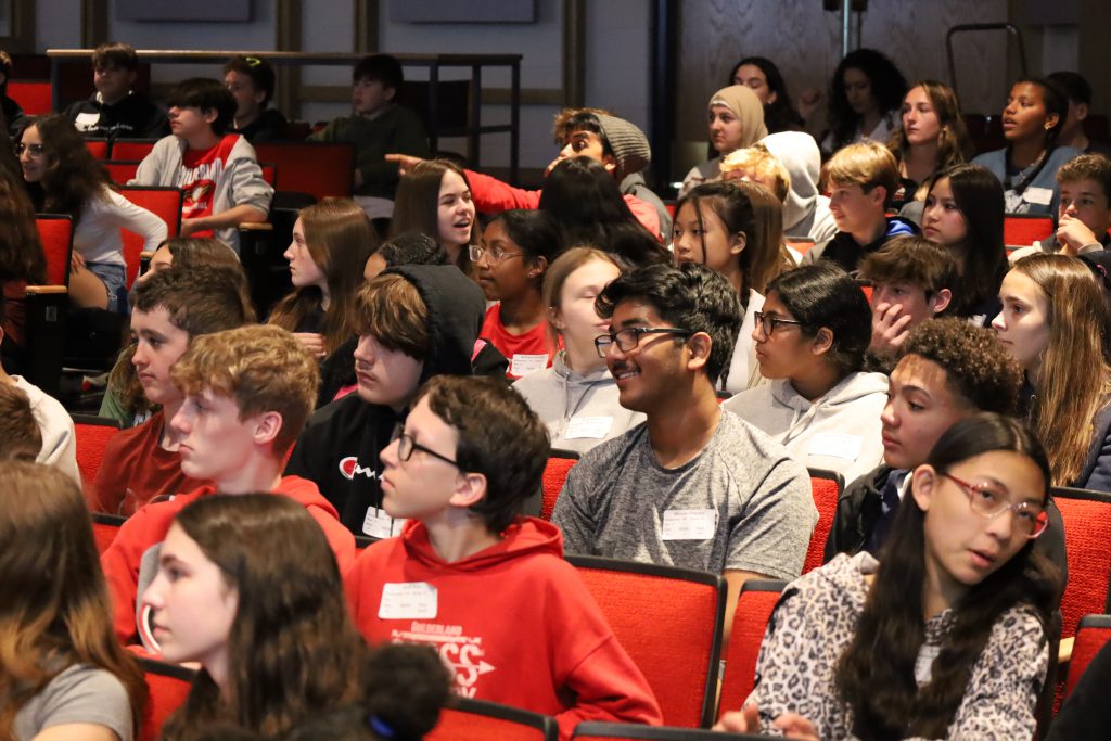 Students are seated in an auditorium. Some are smiling, one is pointing
