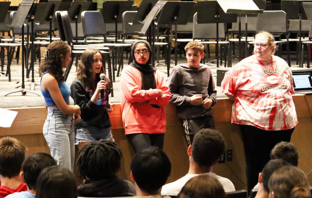 Students and two teachers stand in a line in front of a group of students seated in an auditorium. One is speaking into a microphone.