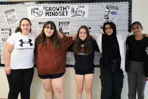 Five students stand arm in arm in front of a bulletin board. The bulletin board reads the power of growth mindset from history.
