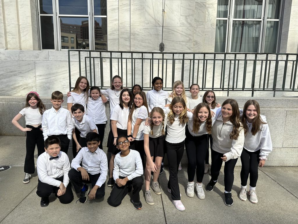 The PBES select chorus is outside, posing for the photo. Some students have their arms around each other, some are kneeling.