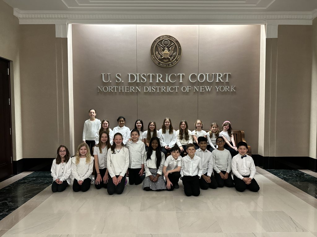 The PBES select chorus, wearing white shirts and black pants, are seated in two rows in front of a wall in the courthouse. On the wall is text that reads: U.S. District Court Northern District of New York