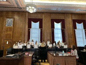 Wide shot of a courtroom in the US federal courthouse. The PBES select chorus is in two rows, singing. In front of them is the chorus director.