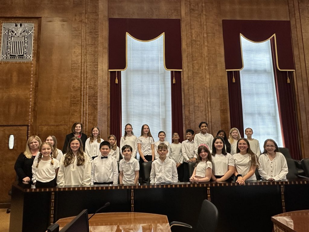 PBES select chorus, wearing white shirts and black pants, stand in two rows in a courtroom. With them are a judge and the chorus director.