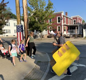 Man speaks through a bullhorn. He is wearing a yellow sign that says Altamont Walking School Bus. The sign is shaped like a school bus and is yellow