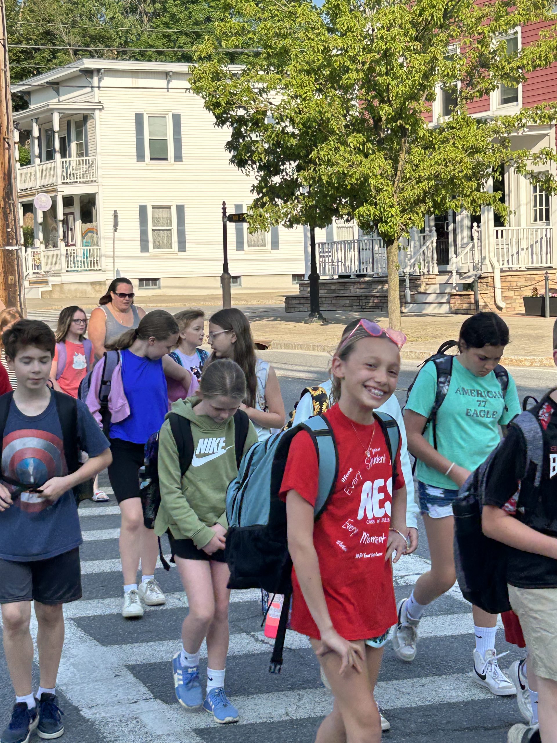 Student smiles at the camera while walking with others to school. The student is wearing a red t-shirt and a backpack.