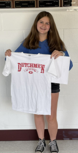 Student stands in front of a white wall holding a white t-shirt with red lettering that reads: Dutchmen of the week