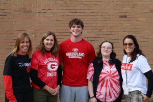 Five people stand in a row, smiling at the camera. Each is wearing a different t-shirt that represents unified athletics at Guilderland High School