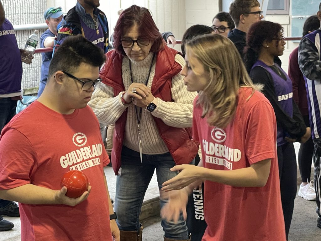 A coach of the unified bocce team speaks to a student who is holding a bocce ball. Another coach looks on.