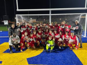 This photo is taken on a yellow and blue field. The team is in the soccer goal. The team is wearing red and white uniforms, holding section champion patches, smiling for the camera. The coaches are standing next to the team. The goalie is wearing neon yellow