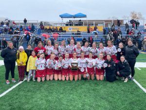 Photo is taken on a field. The field hockey team is wearing white and red uniforms; coaches are wearing black. The team is holding section champion patches, smiling fo r the camera