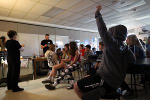 Students are sitting in a classroom. In the foreground a student is raising their hand. In the background an adult is standing, holding an object in his hands that he is looking at.