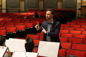 A man conducts a band. He is in an auditorium with rows of red seats behind him and front of him are students sitting with sheets of music in front of them.