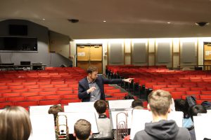 A man conducts a band. He is in an auditorium with rows of red seats behind him and front of him are students sitting with sheets of music in front of them. He is pointing to his right
