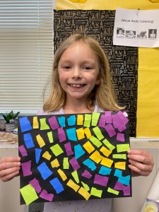 Student is smiling holding a piece of black paper with purple, yellow, blue and green squares glued to it in a curving pattern