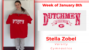 A student stands in front of a white cinder block wall holding a red t-shirt with white text that reads Dutchmen of the Week