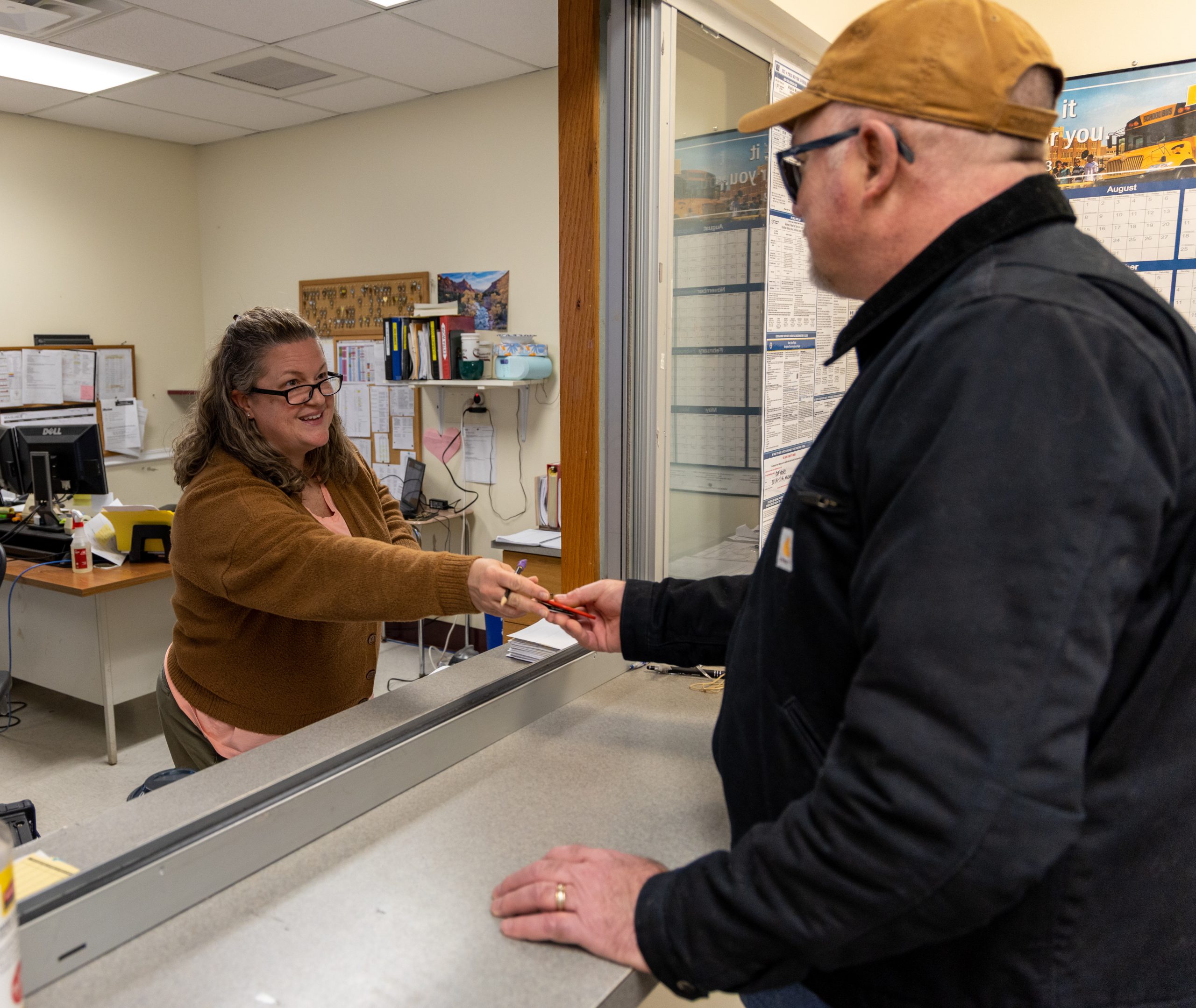 An employee working in an office hands something to another employee, standing at a window