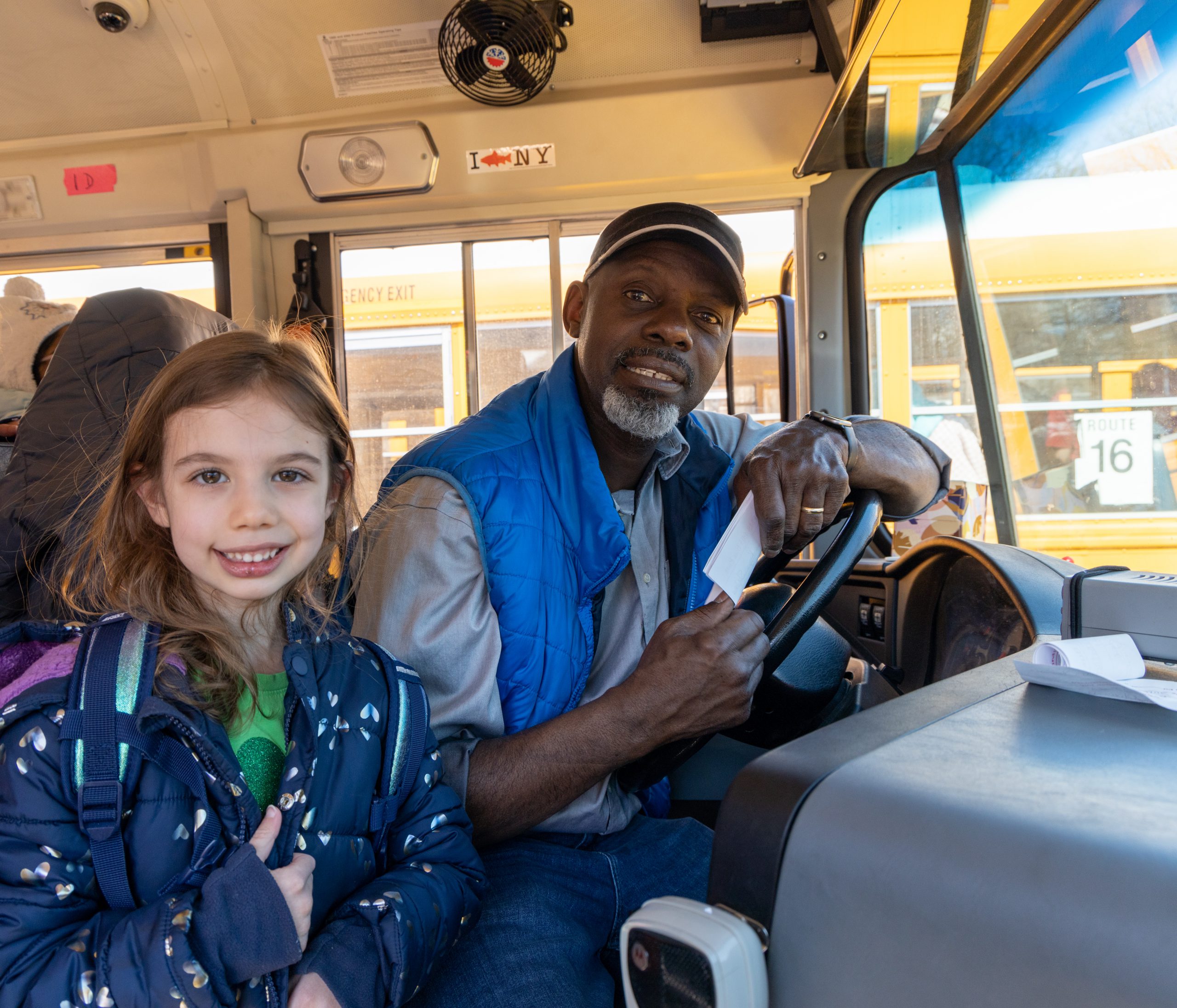 On a school bus, a bus driver and a student standing next to the driver both look at the camera