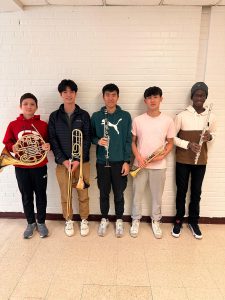 Five students stand against a white wall, each holding a different musical instrument