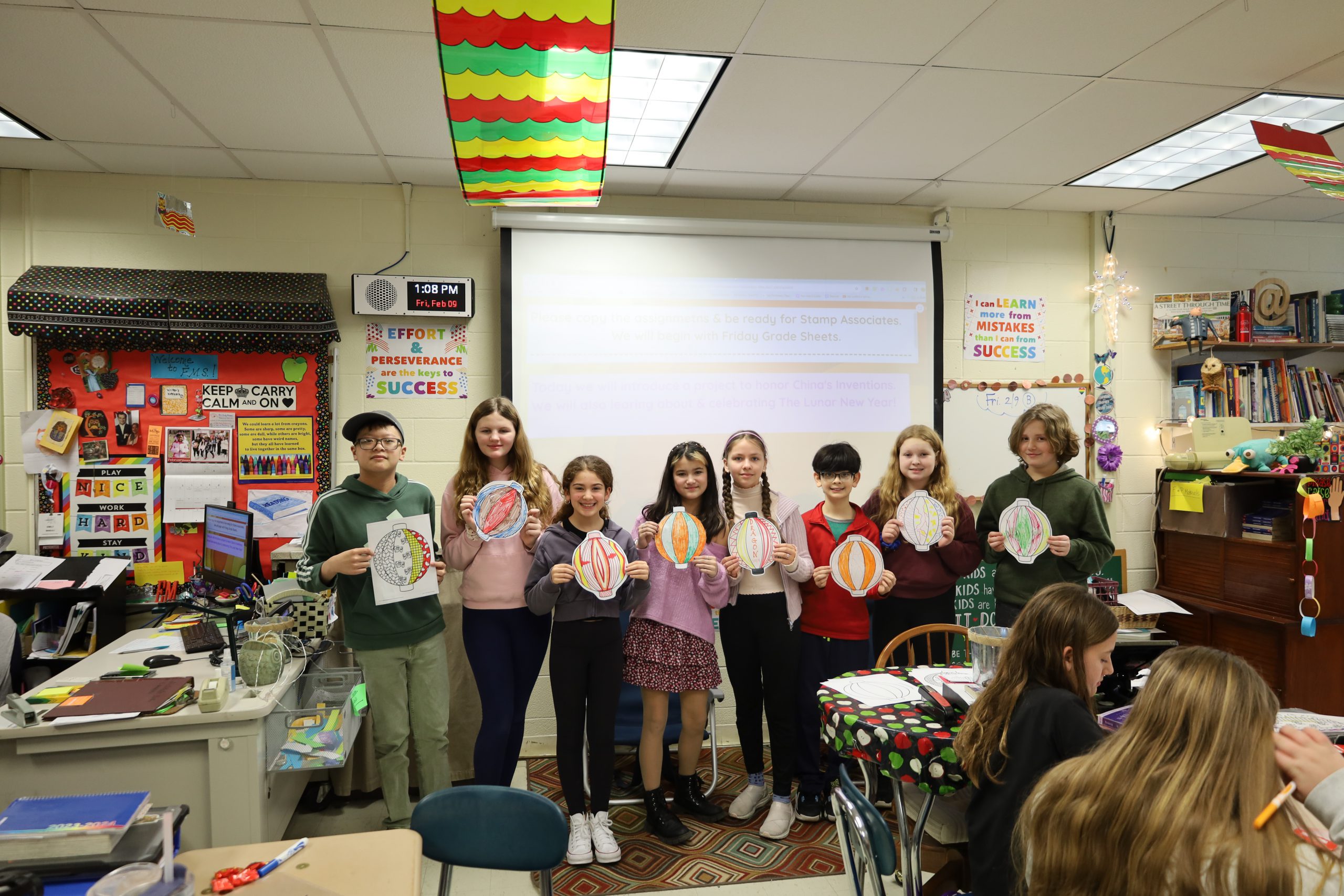 Eight students stand in a line in a classroom holding pictures of lanterns they colored