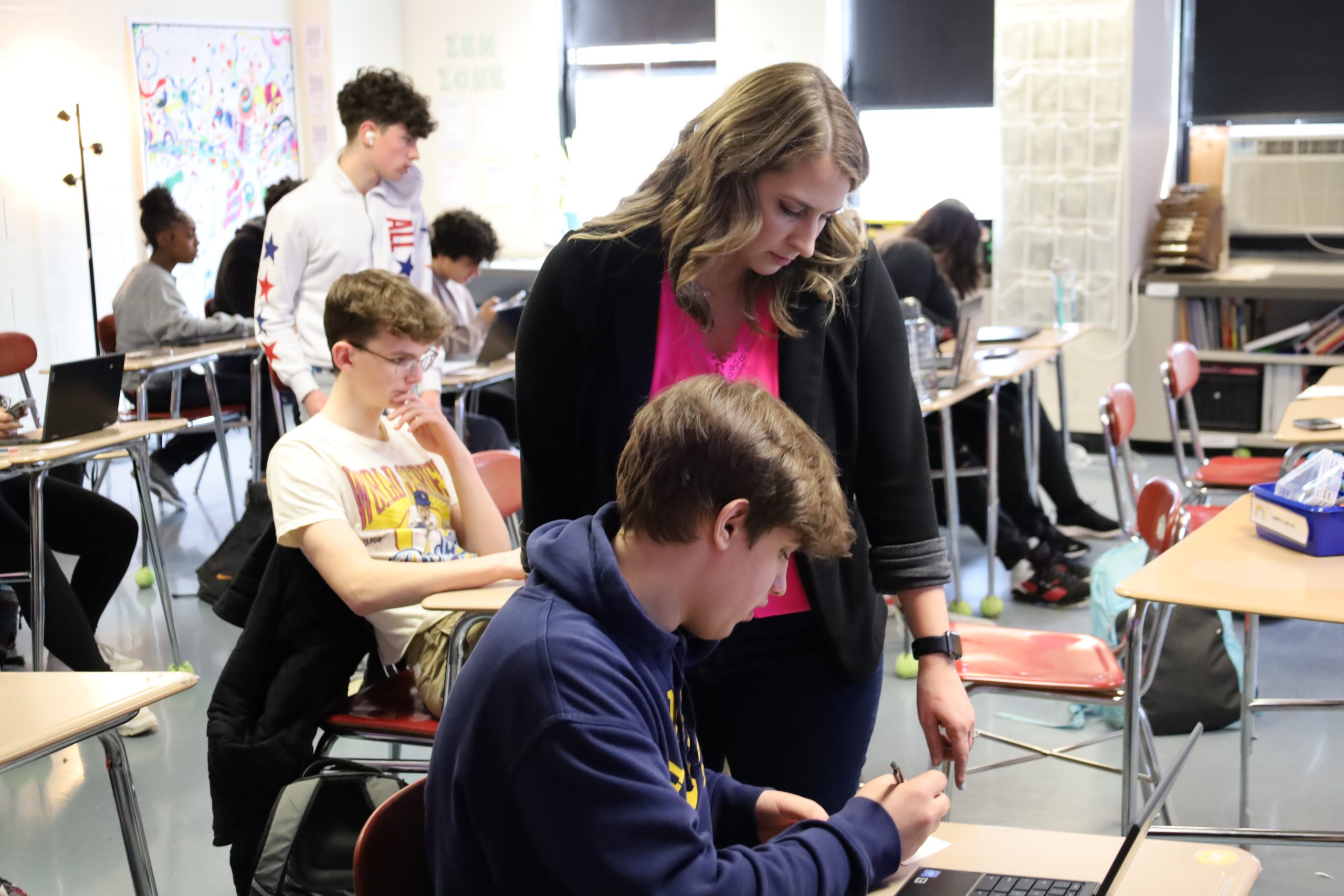 A teacher stands next to a high school student who is sitting at a desk