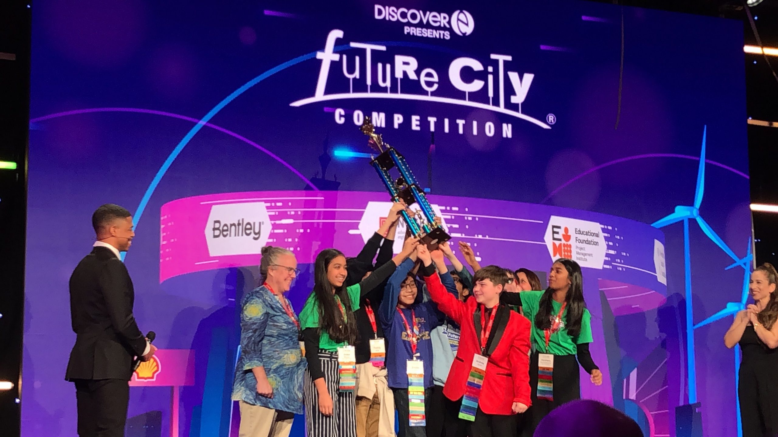 A group of FMS students and a teacher stand on a stage, holding up a trophy over their heads