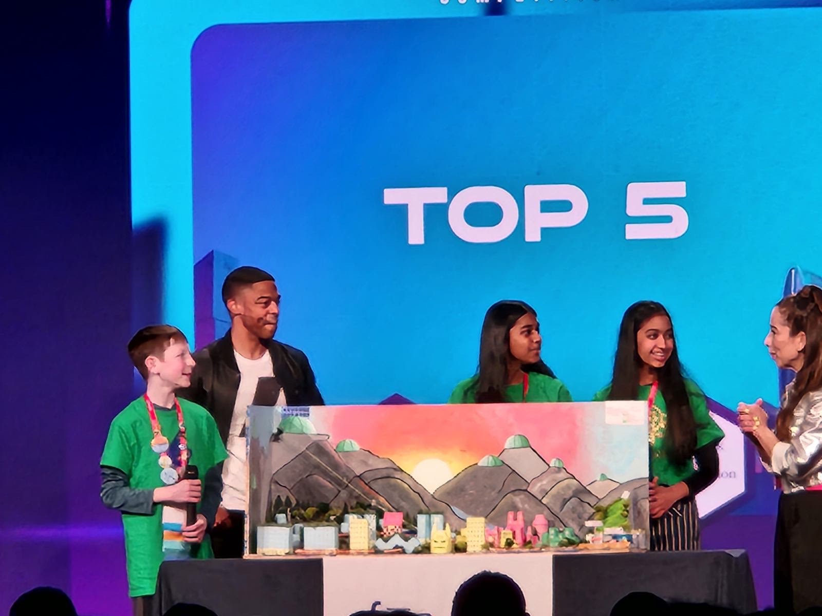 Three students and two adults are on a stage. The backdrop behind them says Top 5. In front of the students is a model of a future city