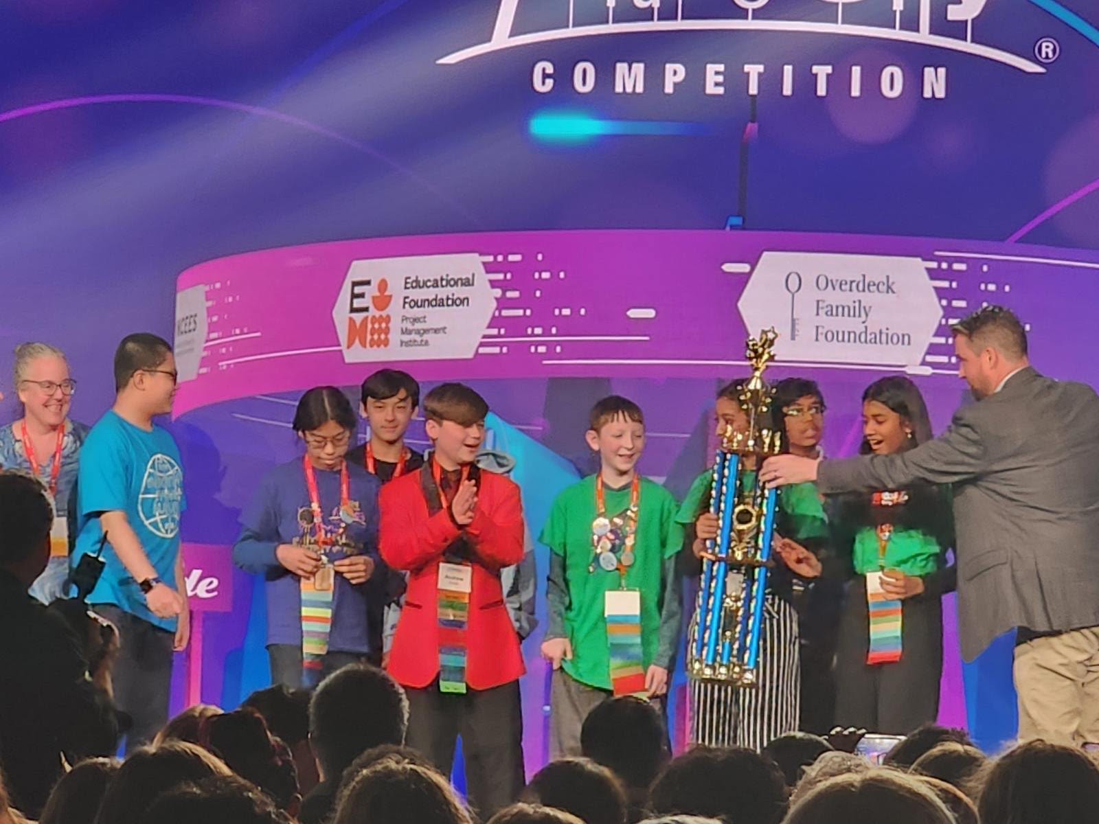 A group of students are standing on a stage, an adult is handing a large trophy to one of the students