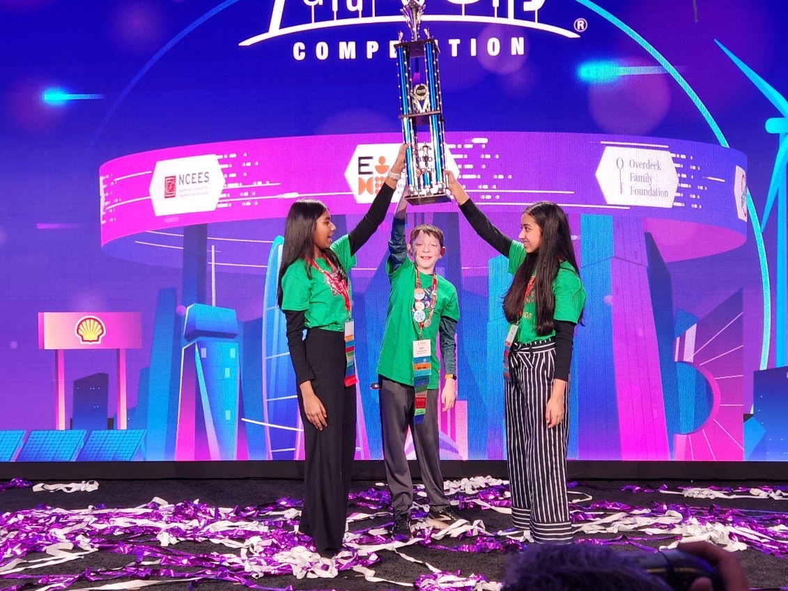 Three students wearing green t-shirts stand on a stage with streamers strewn on it. They are holding a trophy over their heads