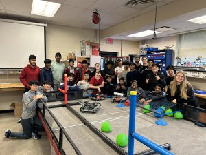 A group of students are in a classroom, gathered around a large table. A few robot-like devices are on the table. The students are smiling at the camera