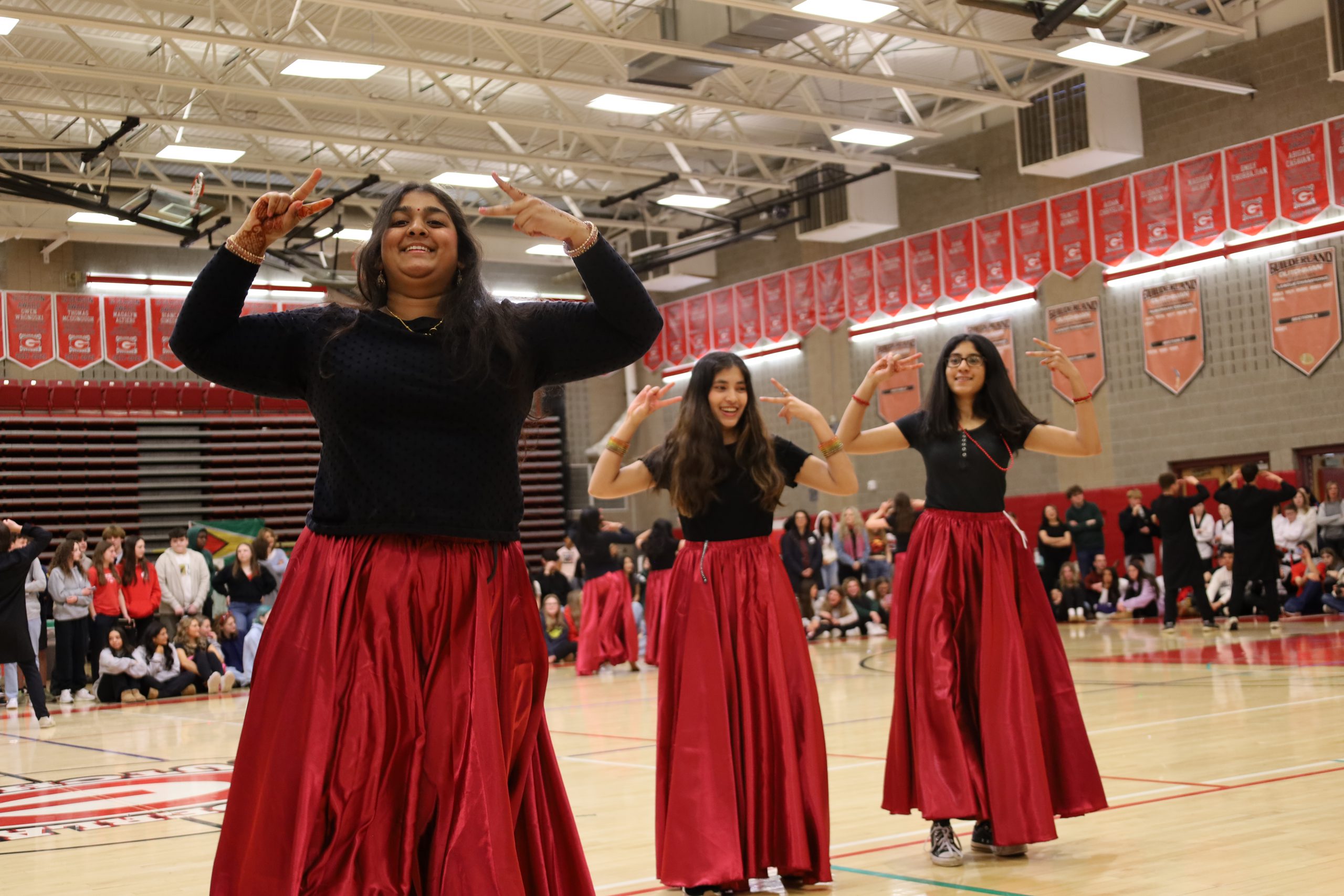 Three students dance in a gym. Their hands are raised, they are wearing black shirts with long red skirts