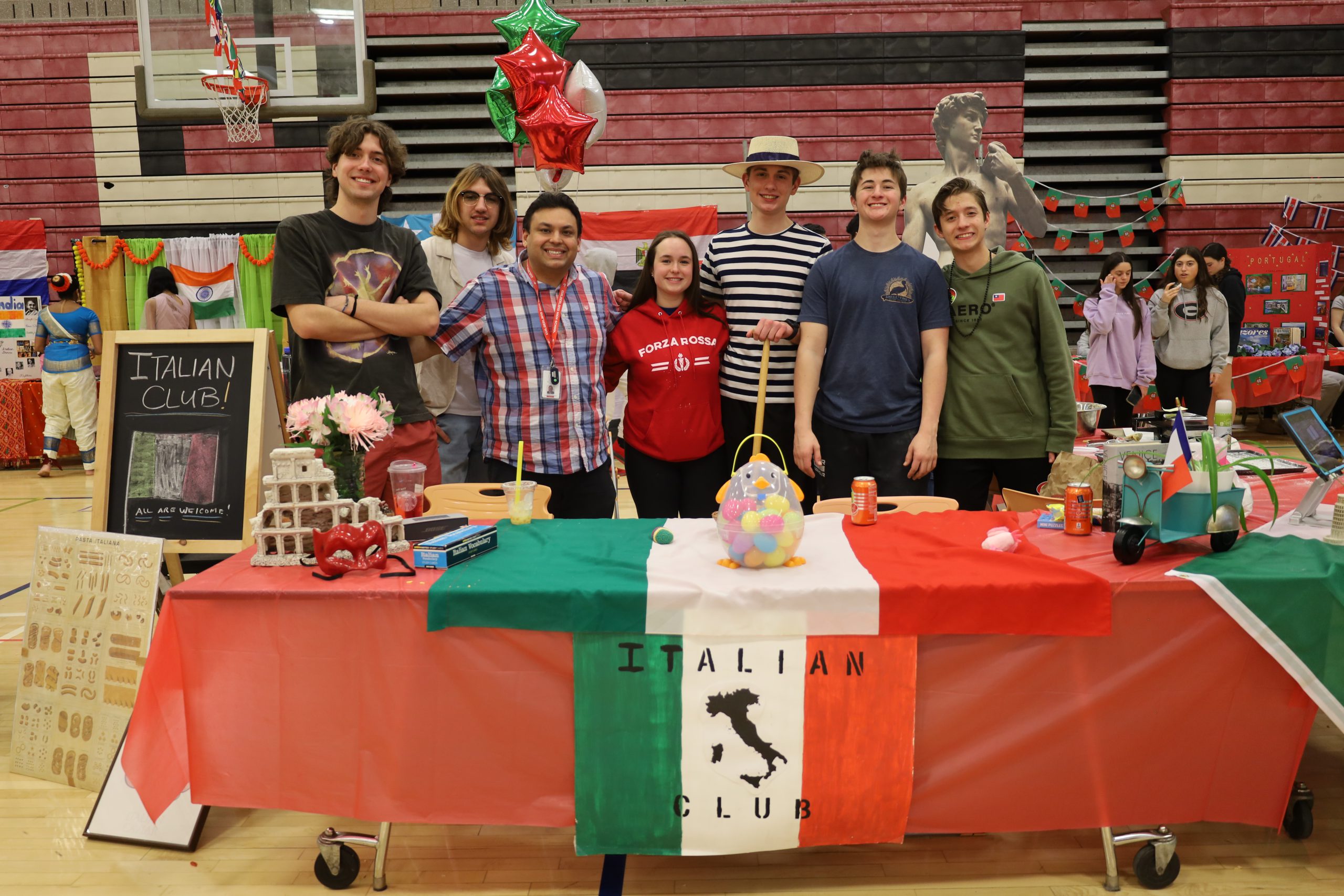 A group of students stand in a gymnasium behind a table with a red white and green tablecloth. A sign reads: Italian Club. The students are smiling. One is wearing a striped shirt and straw hat
