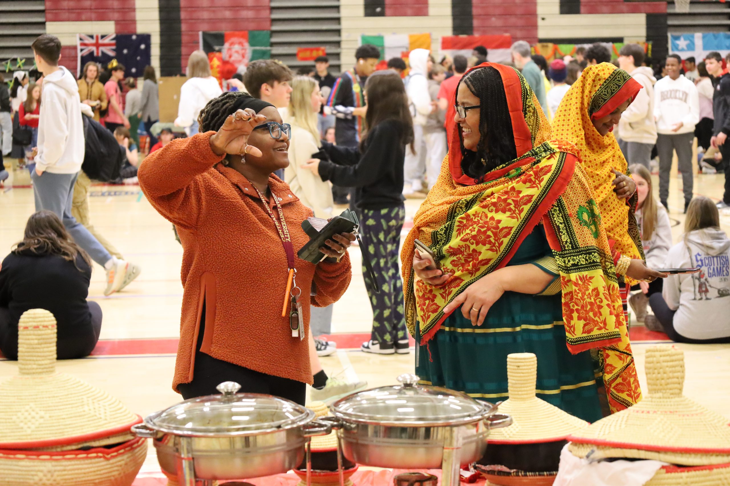 Two people speak in a gymnasium. They are standing next to a table that has straw baskets and food serving dishes. One is wearing traditional clothing and a scarf wrapped around her head