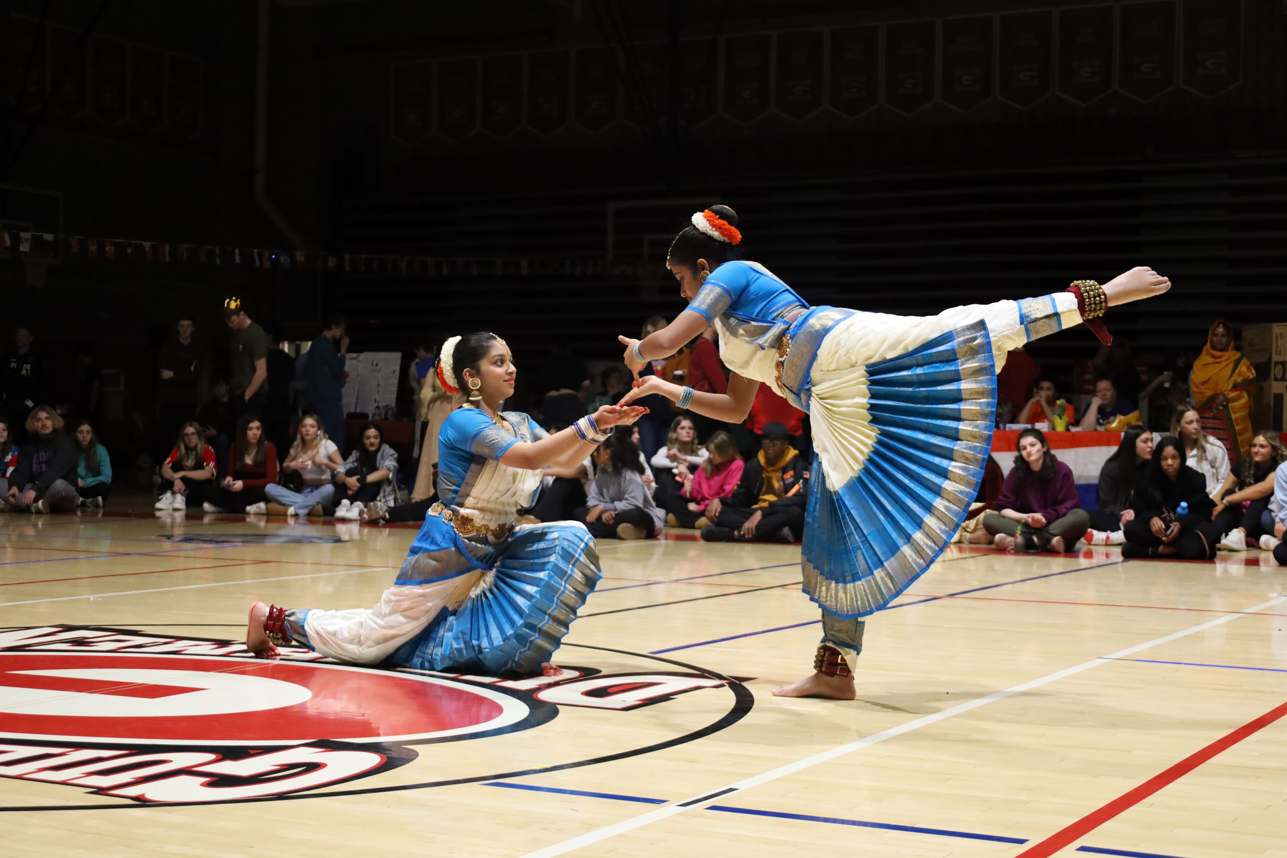 Two students are wearing traditional blue and white clothing. They are dancing. One is kneeling and the other is holding her hand, with her leg extended. They are barefoot.