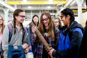 A group of students are in a factory on a tour. They are wearing safety glasses. Two students are holding a large piece of weaponry that is made in the factory
