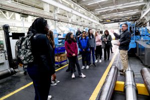 A group of students are standing in a factory. They are on a tour; the tour leader is speaking