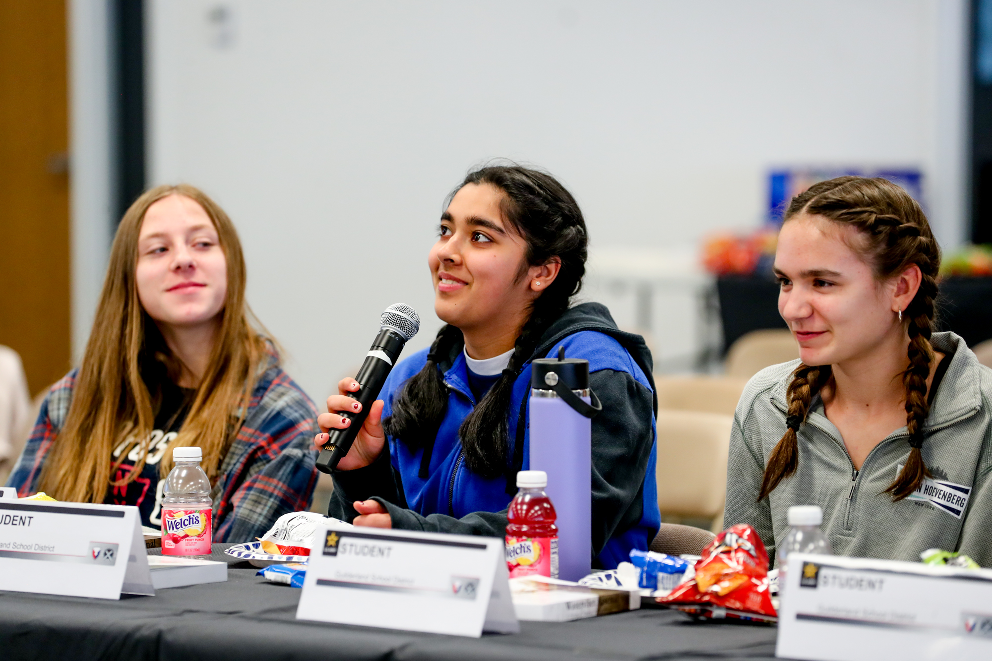 Three GHS students are seated at a table with name plates in front of them. The one in the middle is speaking into a microphone. The student to the left is looking at the speaker