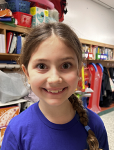 A close-up photo of a student smiling at the camera. Behind her is classroom cubbies