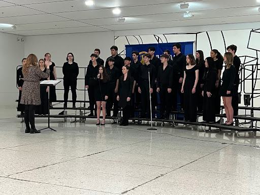 A choir of students wearing black clothing are standing on risers. They are singing. Two microphones are standing in front of them. An adult is standing in front of them, directing them