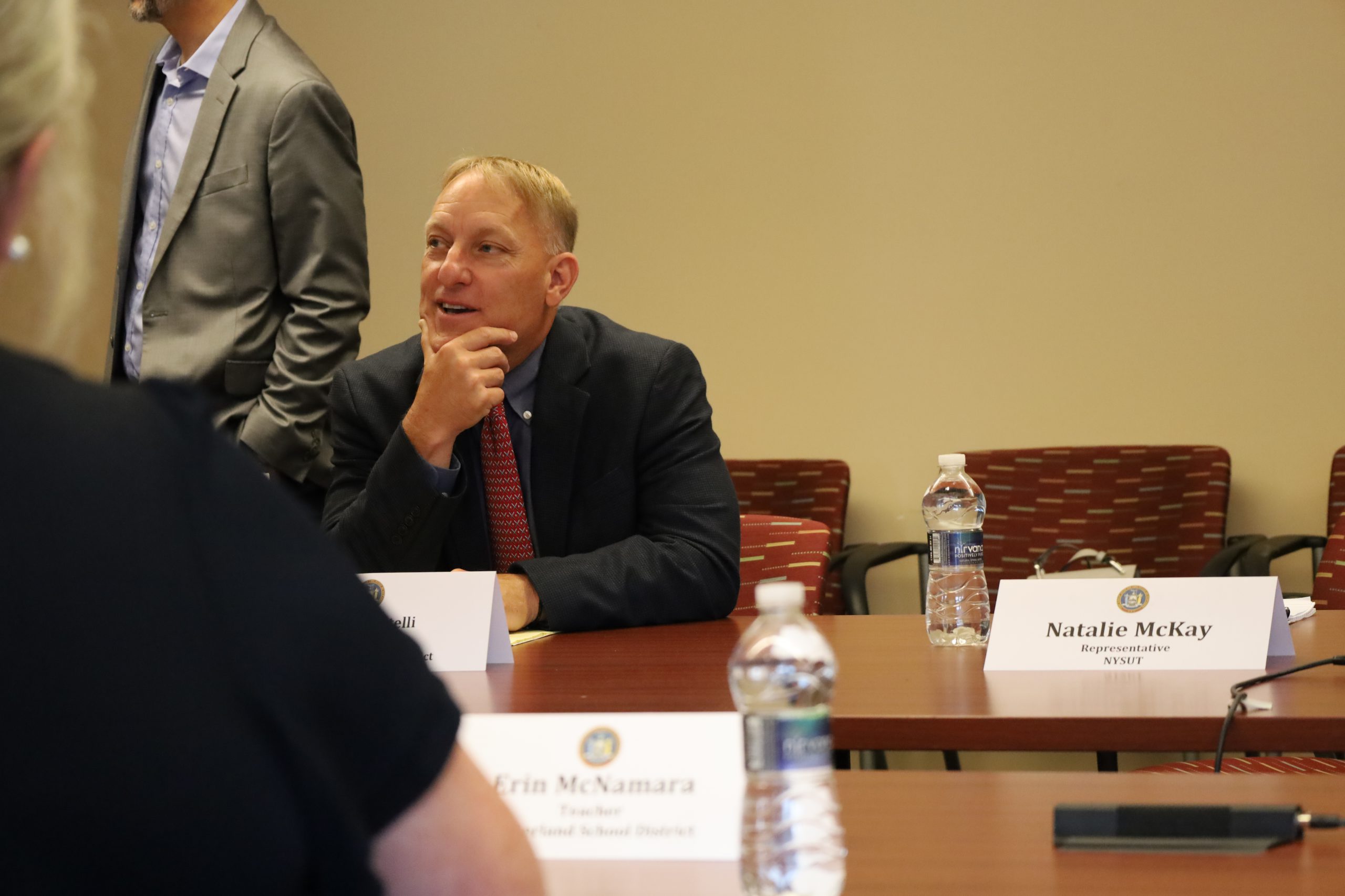 A man is sitting at a conference room table. He is wearing a suit and tie. His hand is to his chin.