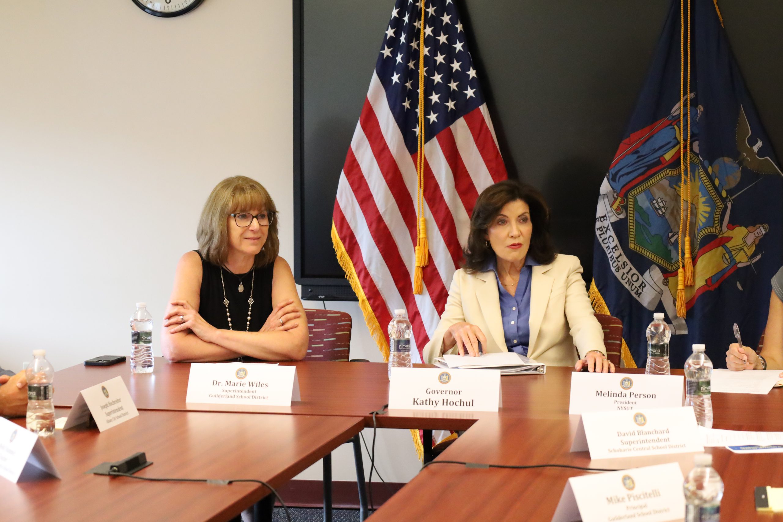 The governor of NY is seated next to the superintendent of schools. at a conference room table. Flags are behind them. They are listening to others speaking