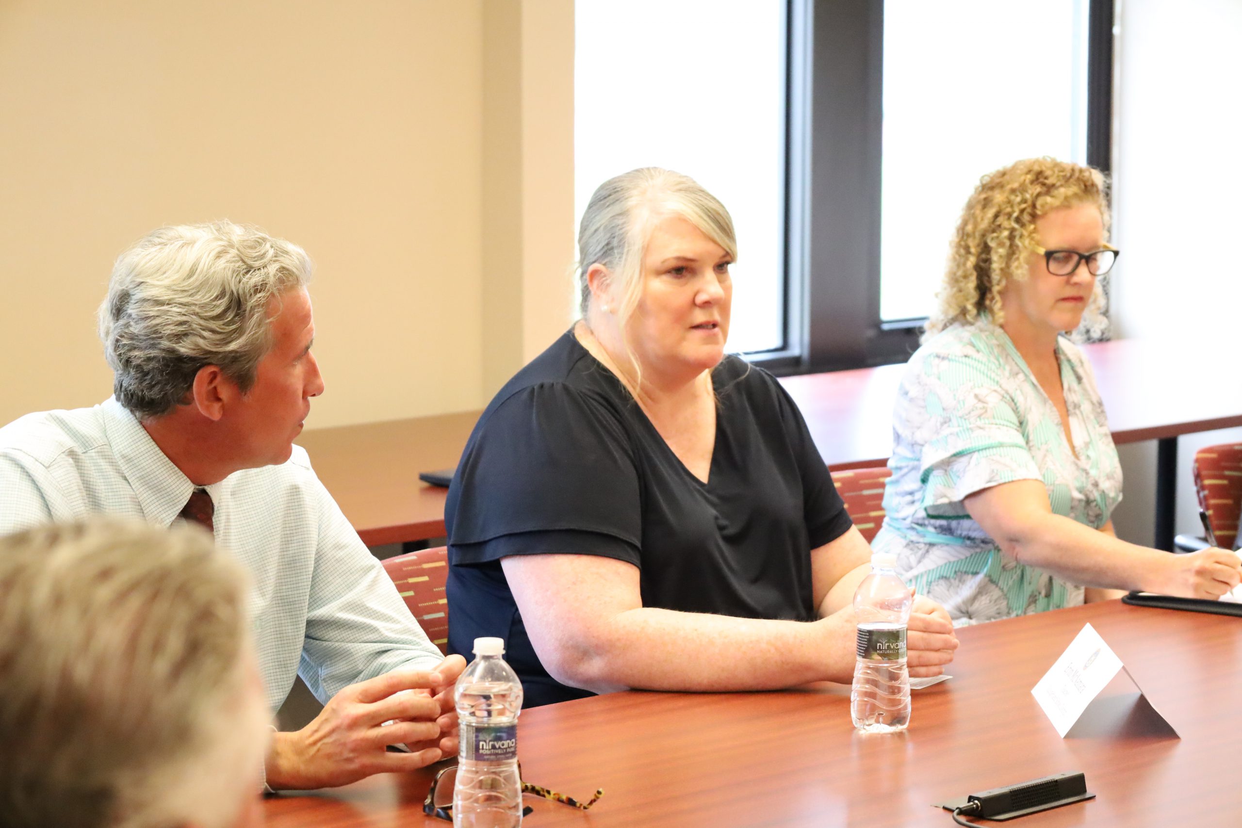 Three people are seated at a conference room table. One is speaking