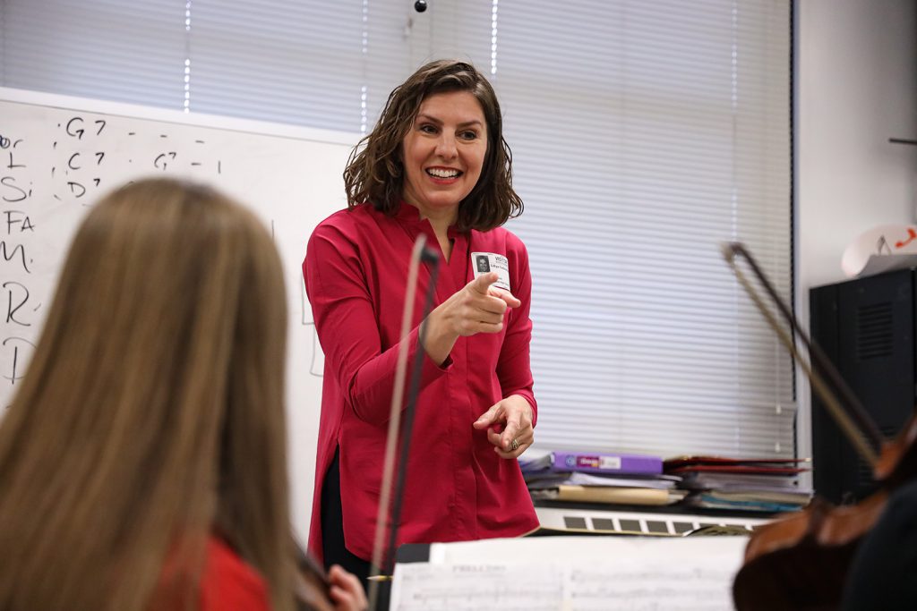 Conductor wearing a red shirt, pointing and smiling. The back of a student's head can be seen to the left of the photo, along with sheet music.