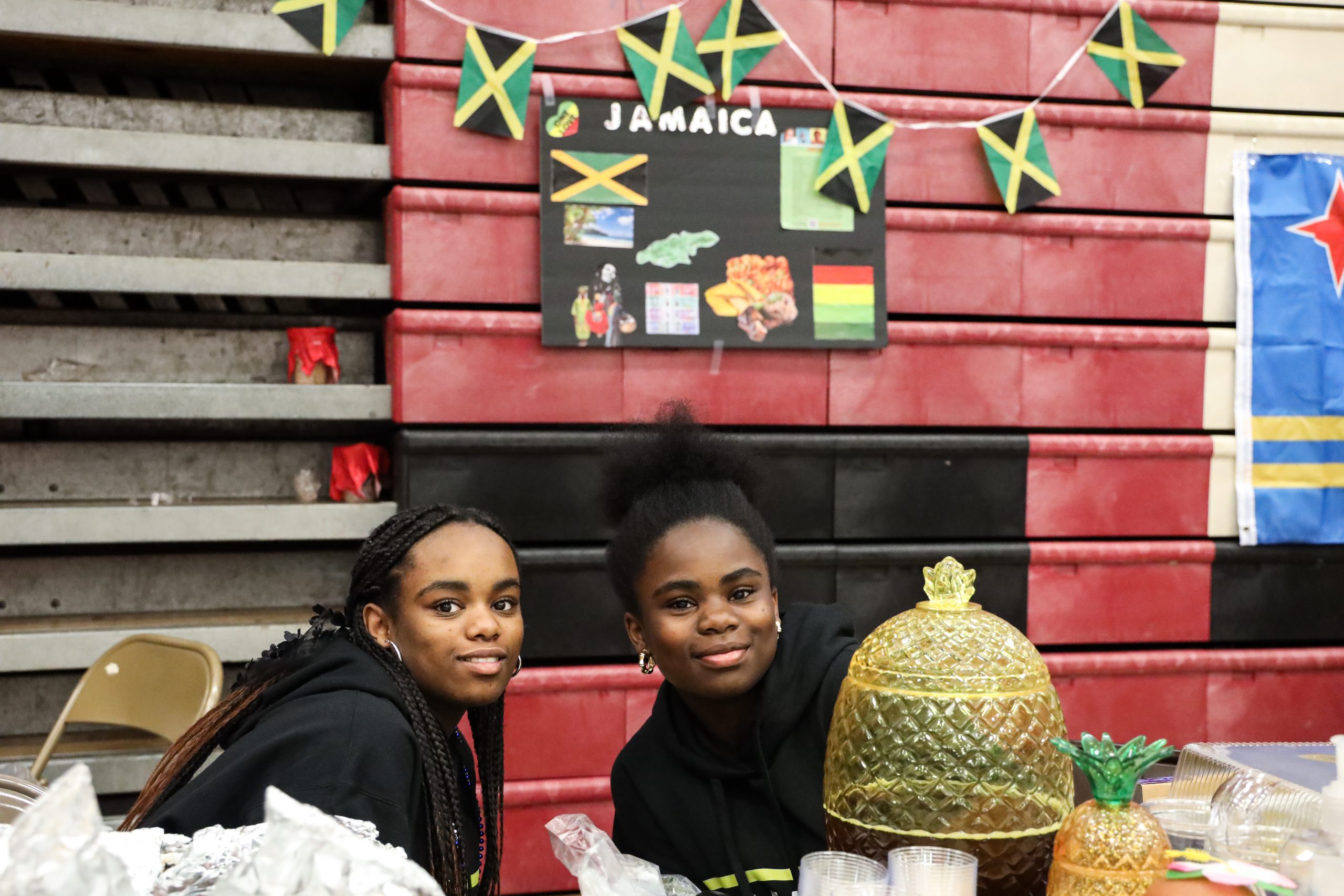 Two students are sitting at a table. They are leaning towards each other as they look at the camera. Behind them is a poster board that says Jamaica and a banner that has small Jamaican flags