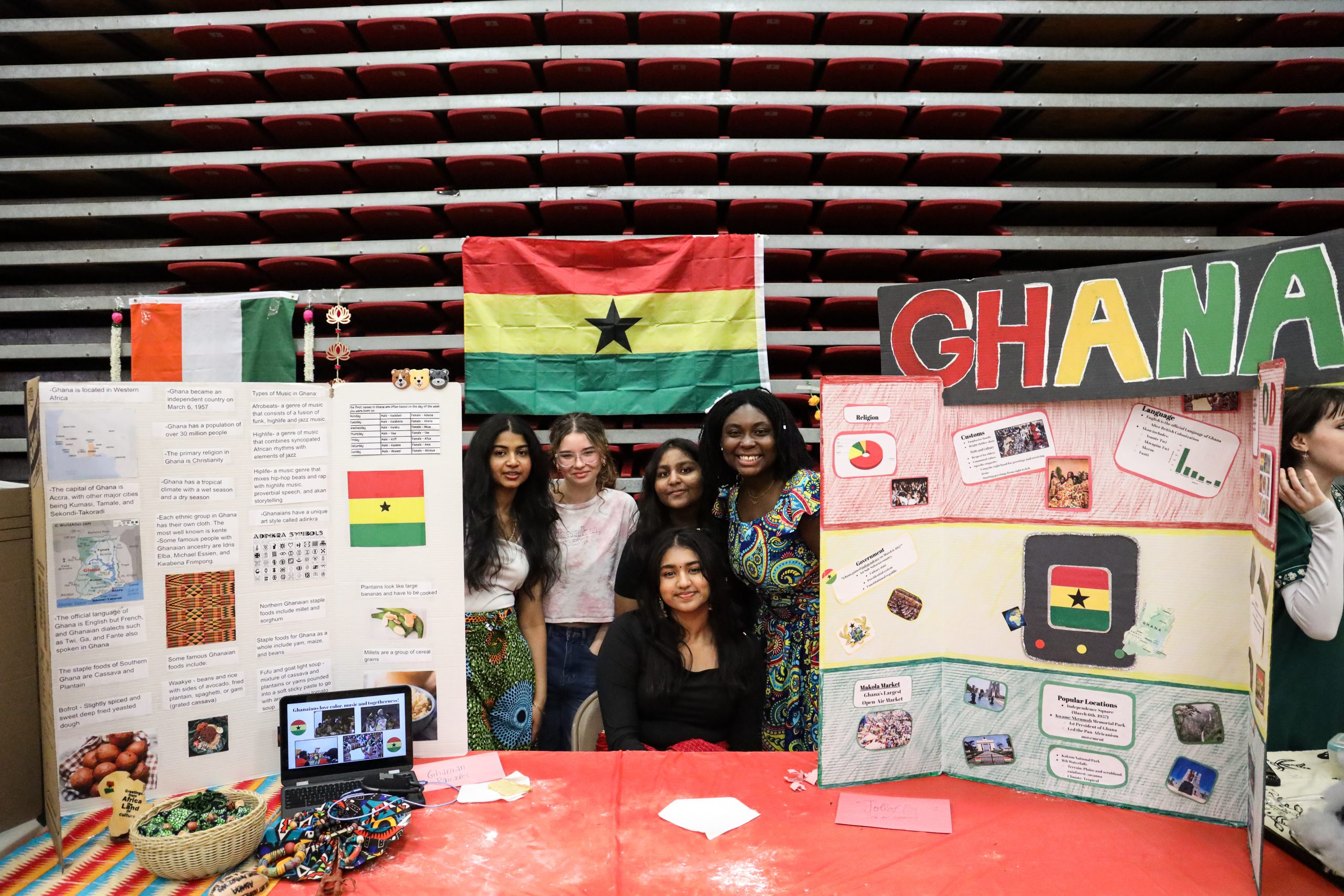 Five students are behind a table. One is sitting the others are standing. On the table are display boards that have information about Ghana